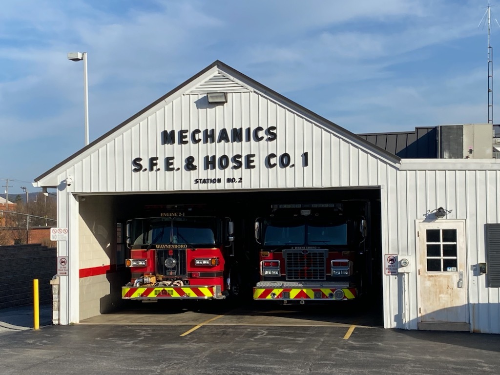 A small fire station with a sign reading “Mechanics S.F.E. & Hose Co. 1” houses two fire trucks in an open garage. The building is white with red accents, and the number “21” is visible on the front of one fire engine. The sky is clear with a few clouds.