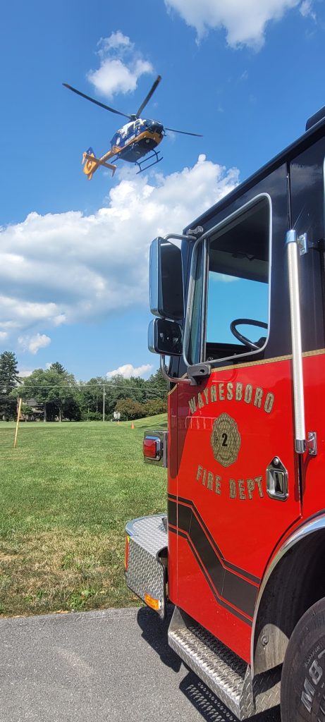 A Waynesboro Fire Department truck is parked on a grassy field under a partly cloudy sky. In the background, a helicopter is flying overhead.