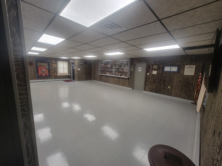 A well-lit, empty room with a tiled floor and wood-paneled walls. The room has a drop ceiling with rectangular light fixtures. Display cases with various items and memorabilia line the far wall, and framed certificates are mounted on the side walls. An American flag stands in the right corner.