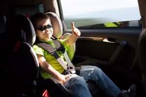 A young child wearing sunglasses and a green shirt is sitting in a car seat, smiling and giving a thumbs-up. The car interior is visible, with sunlight streaming through the windows.