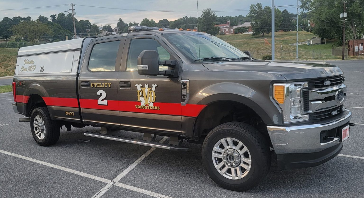 A utility truck with the number "2" and "WRFD" on its side, painted in gray and black with red accents, is parked in an empty parking lot. The truck features a white camper shell and is equipped with a step bar. Trees and buildings are visible in the background.