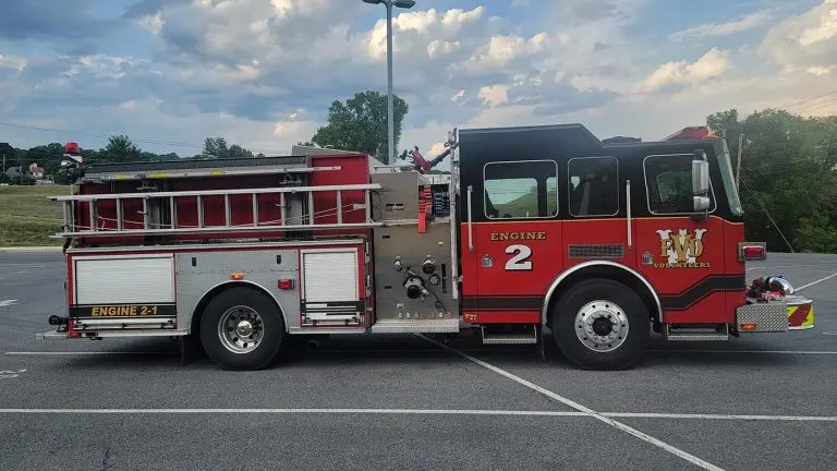 A red and white fire truck labeled "ENGINE 2" and "Engine 2-1" with "LWD Volunteers" on the side is parked in an empty lot. The sky above is partly cloudy, and there are trees and a green hill in the background.