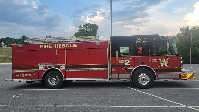 A red fire-rescue truck with the label "FIRE-RESCUE" and the number 2 is parked in a lot. The truck has a logo, emergency lights on top, and various compartments for equipment. The sky behind is partly cloudy.
