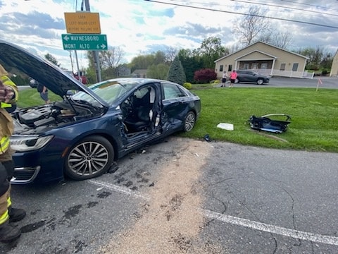 A damaged dark blue sedan with its hood and a side door open is at the side of a road. A detached car part lies nearby on the grass. A firefighter and other individuals are at the scene. A directional road sign and a building with a few people near it are in the background.