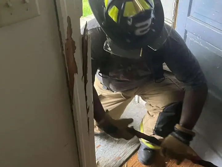 A firefighter in full gear uses a tool to break down a door, causing wood to splinter. Another firefighter stands in the background near an open door, observing the situation. They appear to be performing a rescue or entry operation inside a building.