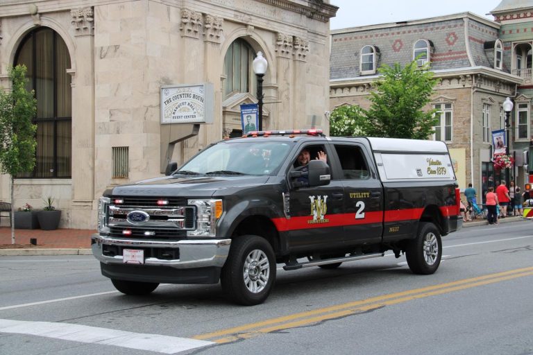 A black and white utility vehicle with "MW" and "2" markings on the side is driving down a street during a parade. The vehicle has red accents and a person inside waving through the open window. Historic buildings are visible in the background.