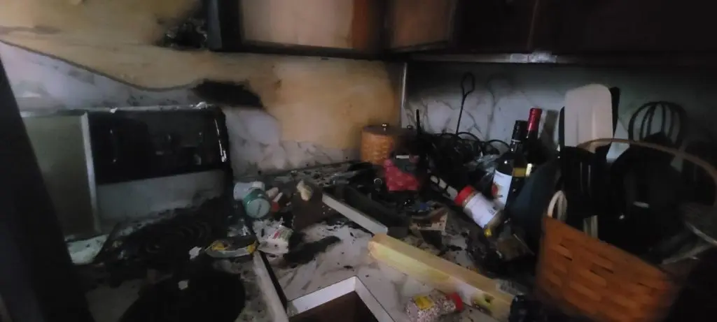 The image depicts a kitchen that has suffered significant fire damage. The walls are blackened with soot, the stovetop appears burned, and there is debris and scattered items, including kitchen utensils and a wine bottle, covering the countertops.