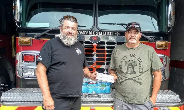 Two men stand in front of a fire truck. The man on the left has a beard and is wearing a black t-shirt. The man on the right is wearing a green t-shirt with a "Liberty or Death" graphic. They are holding a check and standing in front of a pack of bottled water.