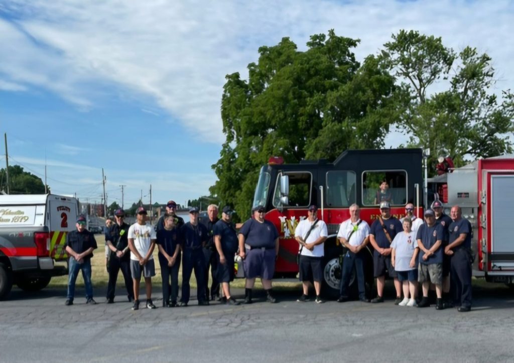 A group of people stands in front of a fire truck on a sunny day. Some are wearing uniforms, likely firefighters, while others are in casual attire. The backdrop includes a green tree and a bright blue sky with scattered clouds.