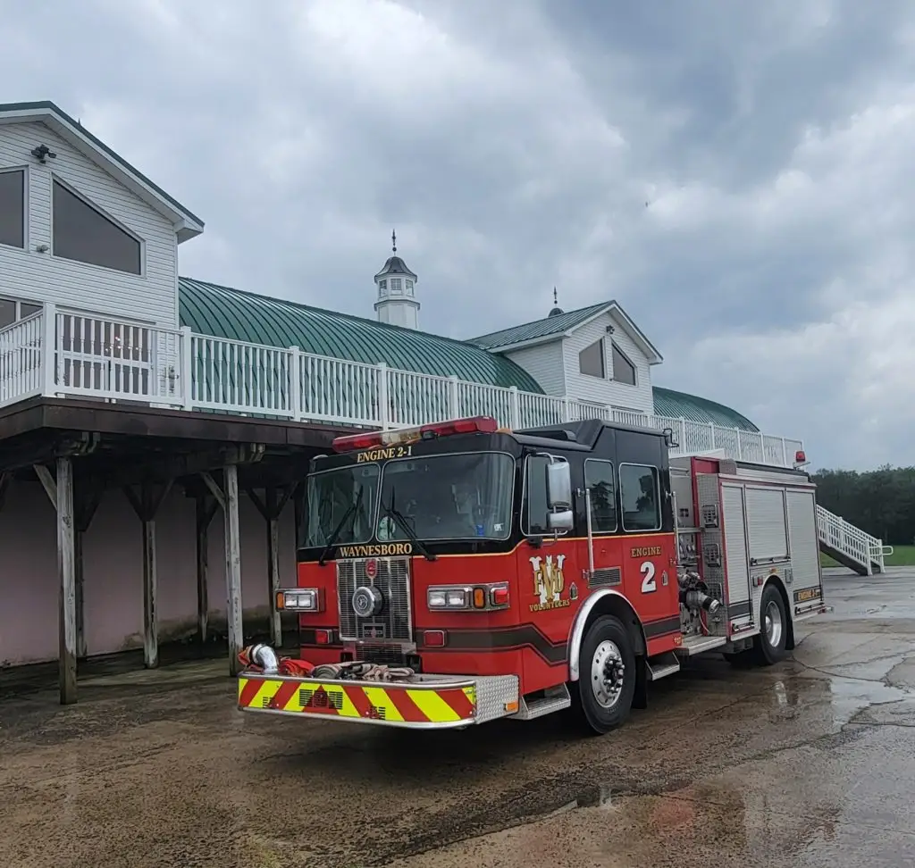A red fire truck labeled "Waynesboro Engine 2" is parked on a wet pavement in front of a large white building with a green roof and a small cupola. The sky is overcast.
