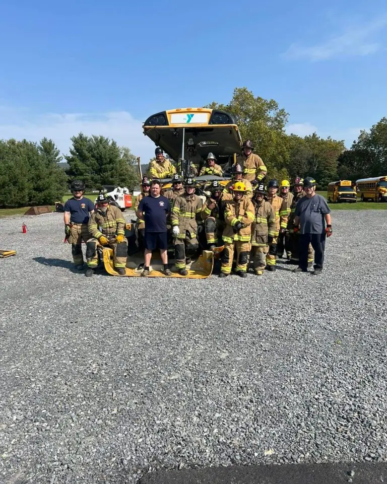 A group of firefighters in full gear stands in front of a fire truck on a gravel lot, posing for a photo. Two people in casual clothes are among the group. Trees and parked vehicles are visible in the background under a clear blue sky.