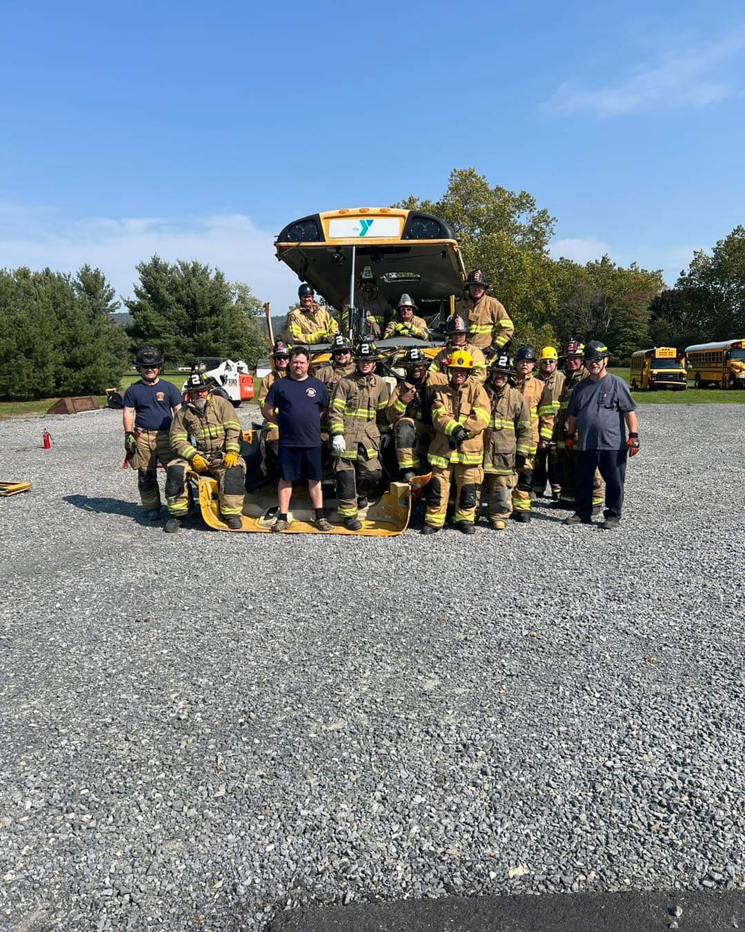 A group of firefighters in full gear stands in front of a fire truck on a gravel lot, posing for a photo. Two people in casual clothes are among the group. Trees and parked vehicles are visible in the background under a clear blue sky.