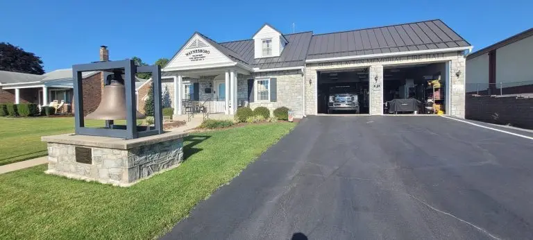 A stone building with a black metal roof, labeled "Martinsburg Volunteer Fire Company." A large bell is displayed on a stone pedestal to the left. The garage doors are open, revealing a fire truck and equipment inside. A neatly manicured lawn lies in front.