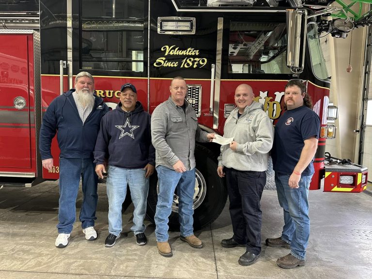 Five men stand in front of a red and black fire truck inside a garage. They are posing for a photo, with one man holding a piece of paper, and all are wearing casual clothes.