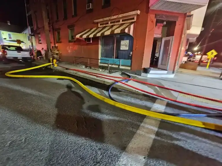 Firefighters and police are on the scene near a brick building with striped awning. Hoses are laid out on the road, and the area is cordoned off. Emergency vehicles are present under street lighting at night.