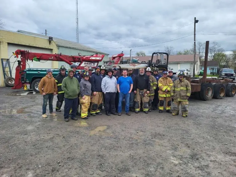 A group of people, including individuals in firefighter uniforms, stands outdoors on a gravel surface in front of various vehicles and equipment. The sky is overcast, and buildings are visible in the background.
