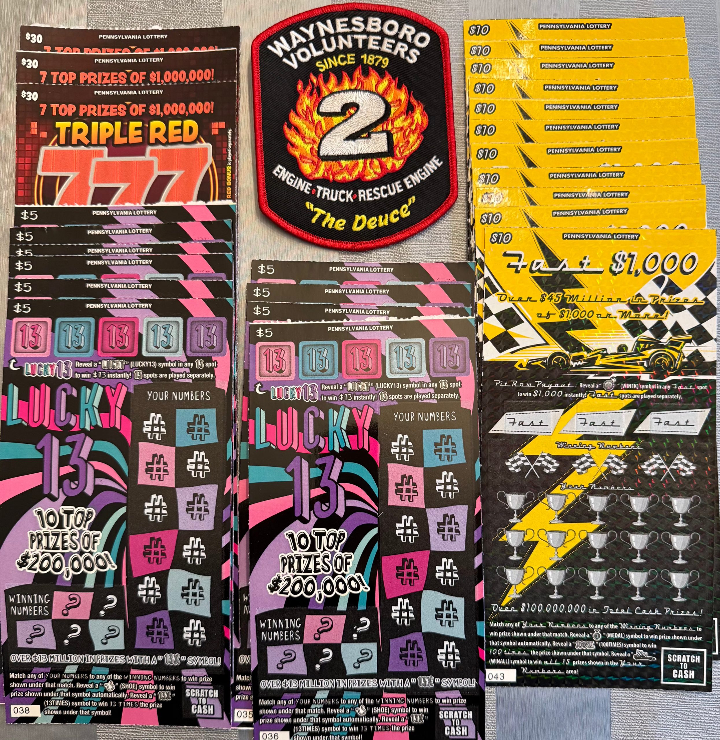 Several colorful lottery scratch-off tickets are arranged on a table next to a Waynesboro Volunteers fire department patch. Some tickets reveal matching numbers and prize amounts, while others remain partially unscratched.