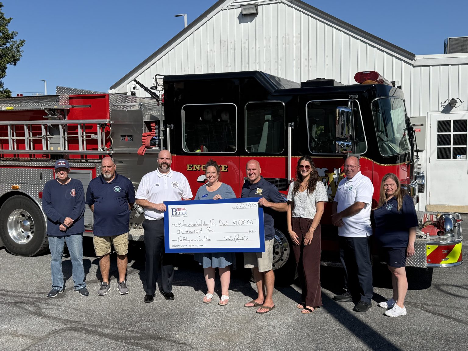 Eight people stand in front of a fire truck, holding an oversized check for $3,000 made out to “Westerly Fire Dept” from “Perryt.” They are smiling and posing for the photo by a white building on a sunny day.