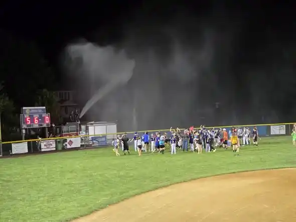 A group of people celebrate on a baseball field at night near a scoreboard showing 5-0, while water sprays over them from a fire truck in the background.