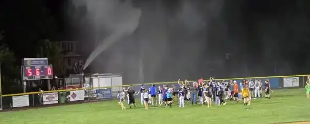 A group of people celebrate on a baseball field at night near a scoreboard showing 5-0, while water sprays over them from a fire truck in the background.